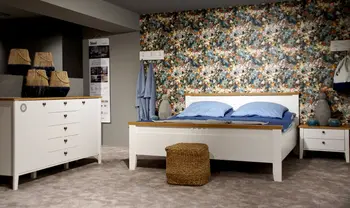 Helles Schlafzimmer im Landhausstil mit weißem Bett, passender Kommode und Nachttisch, blauer Bettwäsche sowie auffälliger Blumen-Tapete als dekorativem Blickfang.